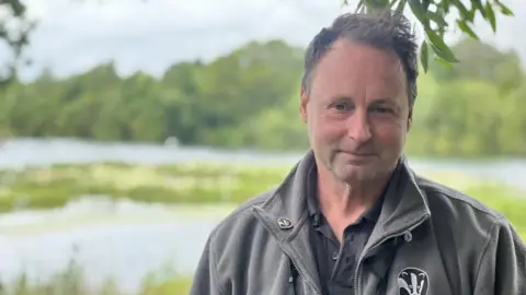 Amber Macey/BBC Warden Grahame Hopwood looking into the camera with grey Lincolnshire Wildlife Trust jacket with the reserve in the background