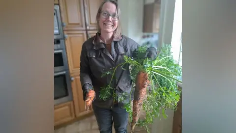 Diana McIntyre standing in her kitchen holding the giant carrot. She is smiling widely and is wearing a khaki waxed jacket and jeans. The carrot is covered in mud and has huge green fronds.
