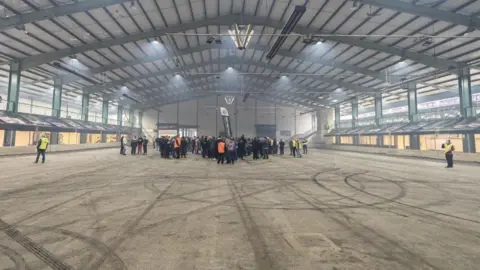 The inside of a large industrial unit, with people stood around in the centre. A series of tyre marks can be seen on the ground.