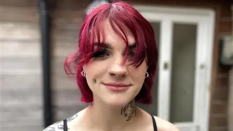 Robby West/BBC Chloe Ponssford standing outside looking directly at the camera and smiling.