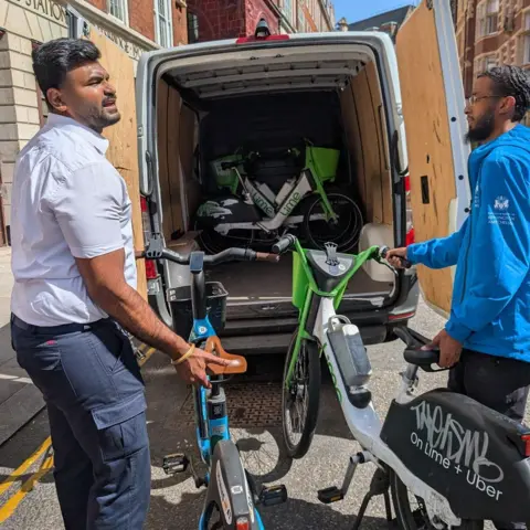 PA Two people load shared bicycles into the back of a white van on a city street, with more bikes visible inside and brick buildings in the background under clear skies.