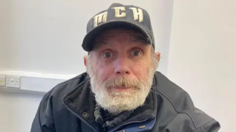 LDRS Gary Morgan, wearing a black jacket and a blue faded denim cap, looks into the camera. The cap has the letters M-C-H on it. He is sat in a room with white walls