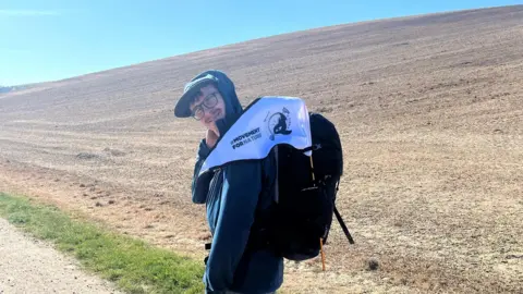 Ben Boxer A man wearing a navy blue hoodie and carrying a backpack with a small white flag on it. The flag has writing on it reading "moving for nature" and a logo of a whale. He is stood on a track with a brown hill behind him.