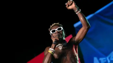 Shatta Wale performing on stage while shirtless and wearing white glasses 