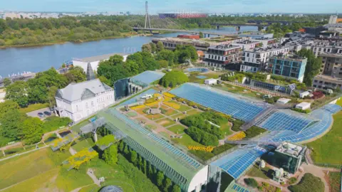 An aerial view of the University of Warsaw with a large river behind it. There is lots of green space and a large white period building in the foreground with modern buildings in the background.