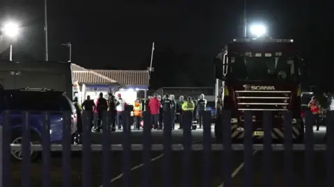 Kevin Saddington/BBC People gathered around a building at night. Many are wearing raincoats and anoraks.  There is a fence in the foreground and a fire engine.