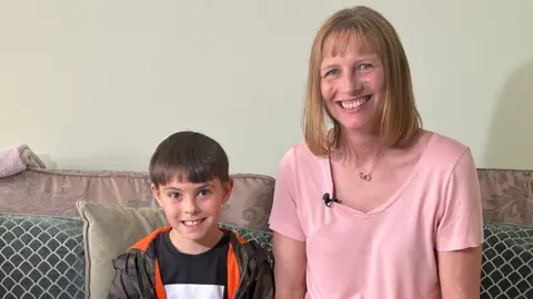 Ruth, wearing a pink top, sits on a sofa next to her eight-year-old son Samuel