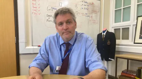 Chas looks into the camera, sitting in the corner of a room with a whiteboard behind him and a mannequin with a school blazer. He wears a blue shirt and tie.