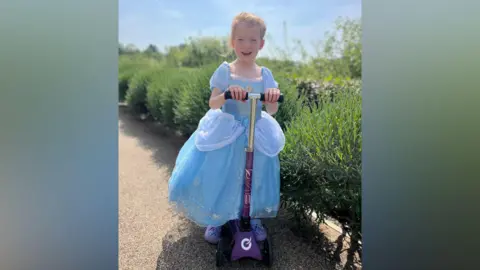 A five-year-old girl in a blue long dress and purple shoes. She is riding her scooter near a park.