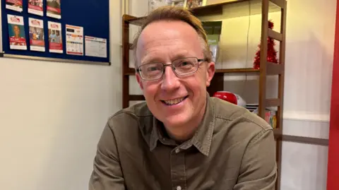 BEN SCHOFIELD/BBC Jon Kuhrt looking directly down the camera, smiling a full smile. He is seated in doors and is wearing a khaki coloured shirt, which is open at the next, and rectangular glasses. He has short hair, that is combed to one side. He is sitting in front of a wooden bookcase and there is a notice board with leaflets pinned to it on the wall on the left of frame.