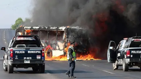 Getty Images An image showing muicipal police trucks next to a burning bus set on fire, and a police man is walking in between. The bus is charred with flames engulfing it from below. The cars are parked on a highway. 