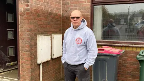 A man with a bald head and wearing dark glasses looks into the camera. He's wearing a grey hoodie with a red logo on the chest. He is standing in front of a brick built house with a window over his left shoulder and a black wheelie bin behind him
