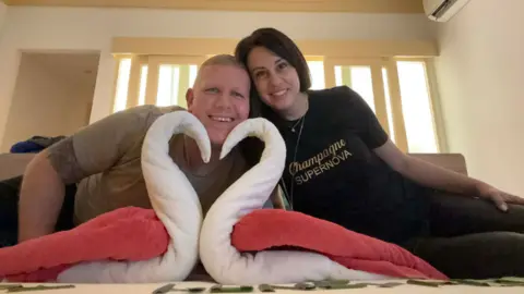 Natalie Chetkowski A smiling man and woman sat on a bed smiling towels made into a heart shape