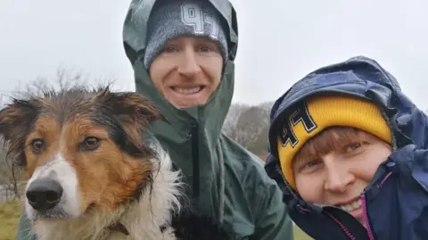 Sam's dog is to the left of the picture, her partner Chris is in the middle and is wearing a khaki green waterproof with the hood up and a woollen hat underneath, Sam is in a navy waterproof with a yellow woollen hat underneath, it is raining, they are out on a hike and are all quite damp.