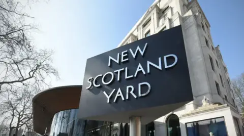 BBC A stock image of the famous triangular New Scotland Yard sign outside the Metropolitan Police headquarters on a sunny day with a blue sky in the background.