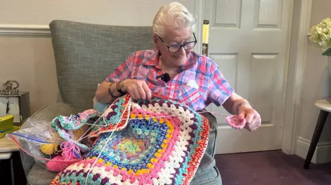 A woman with short white hair and glasses wearing a light ink and blue check short sleeved shirt is sat on a large grey armchair. On her lap is a crocheted blanket, beside her are a few balls of wool and she has a small square of the crocheting in her hands.