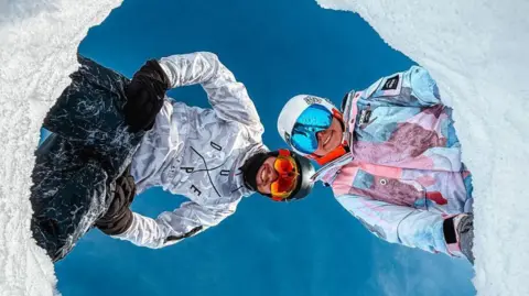 That Travel Couple Hannah Bird and Charlie Camper wearing full snow suits, helmets and ski goggles. They have dug a hole in the snow and placed their phone in there, pointing upwards to capture them looking down against the bright blue sky.