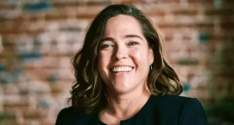 A smiling woman with shoulder-length hair which is brown with caramel highlights. She is wearing a black top. Behind her is an exposed brick wall.