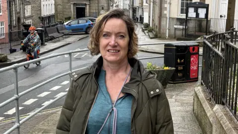 Sinead had light brown shoulder length hair and wears a brown jacket and blue jumper. She stands in Penryn town centre  with a road behind her.