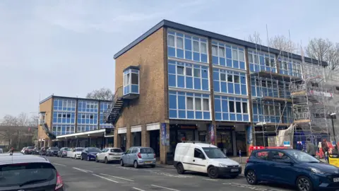 A row of 1960s-style three-storey brick and glass buildings lines a local shopping parade, with a Farmfoods store at ground level. Scaffolding is visible on the right-hand side of the building, and cars are parked along the road in front. The sky is overcast and the street is quiet with a few pedestrians visible in the distance.