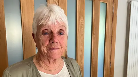 James Grant/BBC A woman in a grey jacket standing in front of a wood and glass door.