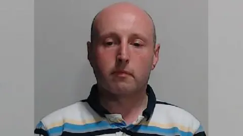 Ross Jameson mugshot. A bald man wearing a blue, white and yellow striped polo-shirt. 