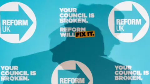 The shadow of Reform leader Nigel Farage is cast onto a Reform UK-branded blue board during a press conference in Doncaster, ahead of May's local and mayoral elections. It reads "Your council is broken, Reform will fix it".