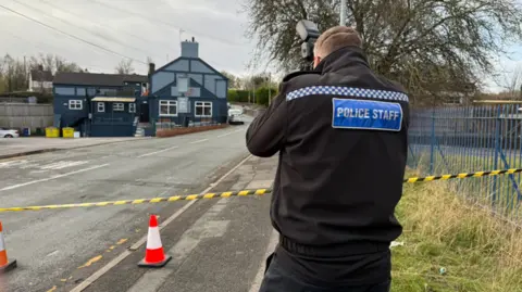 A person in a black jacket with the words police staff on the back is taking pictures from the side of a road. There are cones and yellow and black tape across the road.