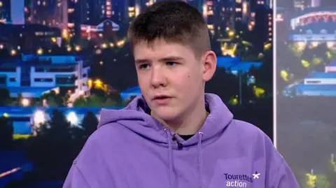 Dexter, 13, with short brown hair wearing a purple hoodie with Tourettes Action logo on it speaking in BBC North West Tonight's TV studio
