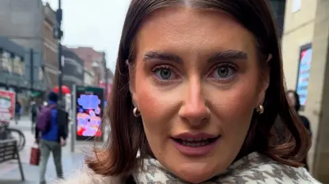 Woman looking at camera. She is wearing a cream and light grey geometric print scarf, has short brown hair and is wearing gold small hoop earrings.