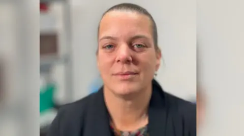 A head and shoulder shot of Emma King, wearing a patterned top underneath a black cardigan. She has blue eyes and dark brown hair which is scraped back into a tight bun, and is looking at the camera with a slight smile.