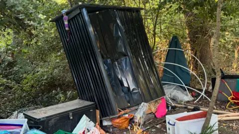 Little Wild Tribe A large black metal cupboard with a big dent in it leans on its side, with various children's items poking out of the bottom. Other play equipment, including hula hoops, are strewn around the site.