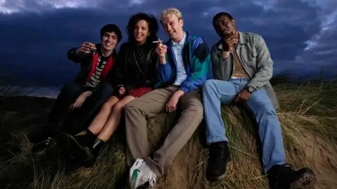 Keeper Pictures and Cyprus Avenue Films Four friends sit smiling and smoking on a grassy mound in a rural field. Clouds roll through a dark sky overhead. 