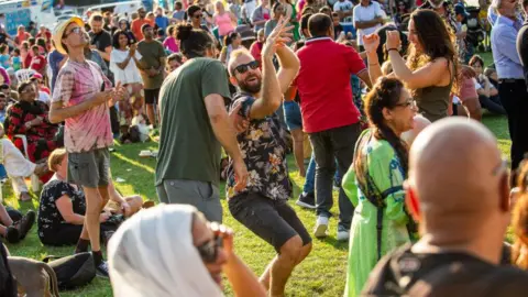 A crowd of people can be seen standing around while some people dance and chat in brightly coloured clothing in a park.