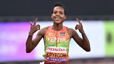 Faith Kipyegon, wearing an orange and green running vest, holds both index finders up towards her face as she smiles in celebration after winning a race