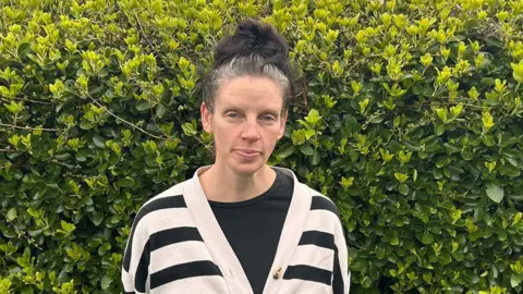 Afton Southern A woman called Afton Southern stands in front of a bush. Her greying black hair is tied into a top knot. She has a black and white stripey cardigan on and a black T-shirt underneath.