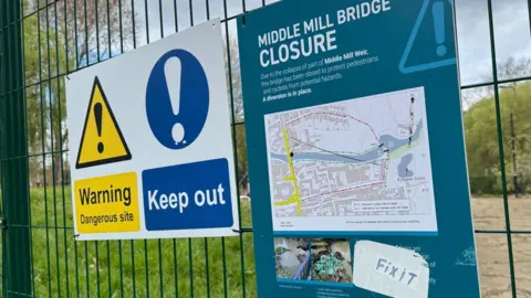 Two signs attached to a green wire fence. On the left is a white rectangular sign with a yellow warning sign above the words "Warning. Dangerous site" and a blue warning sign above the words "Keep out". On the right is a blue sign, about A4 size, with details of a diversion route.