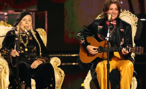 Getty Images Joni Mitchell on stage with Brandi Carlile. Both musicians are sitting on thrones and singing into golden microphones