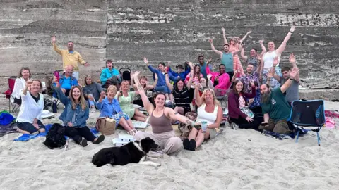 Large group of people seated and standing on a sandy beach in front of a concrete wall, raising their arms toward the camera. A dog lies on the sand in the foreground, with picnic items and bags scattered around.