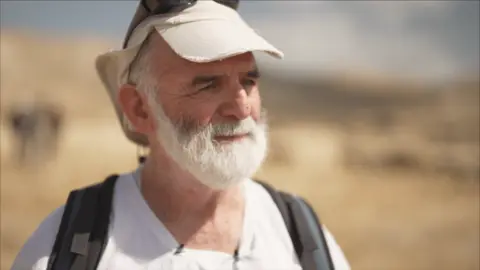 Gil Alexander standing in the sun, shown from the shoulders up. He wears a cap on his head and looks off-camera.