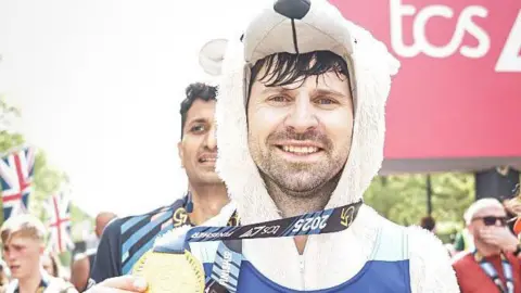 Sportograf A man dressed in a polar bear onesie smiles as he holds a medal, which is around his neck, in his right hand. He is standing at the end of a marathon - other runners can be seen behind him.