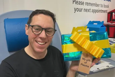 NHS Blood and Transplant A man with dark hair, wearing black glasses and a black t-shirt, sits in a blood transplant chair while holding a blue parcel wrapped to look like a Christmas present. It has a brown gift tag on which reads: "My gift of Plasma".
