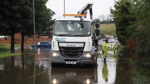 Simon Burns A wide view of the flooded Dublin Road in Enniskillen. A large white truck with a Greentown Drainage Services sign is parked in the middle of the road. Two men in yellow high-viz suits and wellies are standing ankle-deep in the water. IN the distance, a man leans into a blue hatchback car which is submerged up to its bumper in the floodwater.