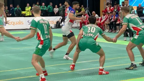 Three men in green kit form a line on a green court. Opposition players in red, blue and white are nearby.
