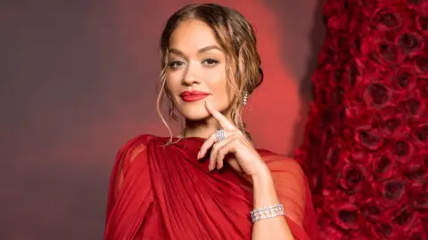 Getty Images Rita Ora poses on a red carpet in a red dress with diamond jewellery 