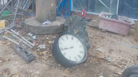 A statue with a clock attached to it lays on the floor, damaged, inside a construction site. A plinth can be seen behind the clock, where the clock had been before it was damaged. 