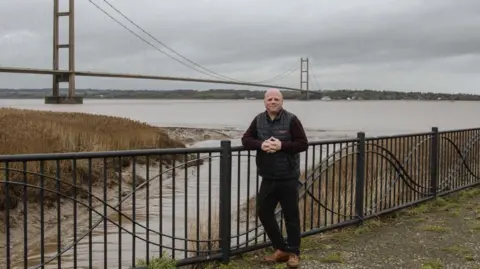 A man standing against some railings with the Humber Bridge in the background. He is wearing dark clothing and has short hair.
