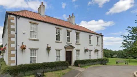 Shotton Hall, where Peterlee Town Council is based, is a two-storey white building surrounded by hedges and grassland. Flower pots are hanging between the windows on the ground floor. There is a wooden bench near the building.
