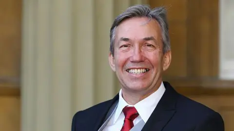 Nick Webborn looks at the camera and smiles. He is wearing a dark suit, white shirt and a red tie.