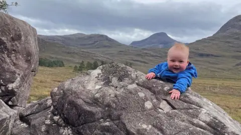 Morag Skelton Hamish being held on top of a rock, with mountains in the background.